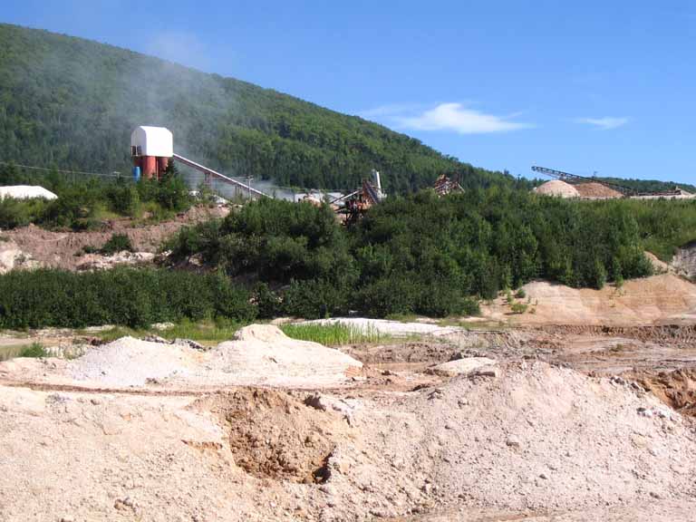 Photographie couleur d’une mine avec une roche blanche au premier plan, un bâtiment et des collines vertes à l’arrière-plan