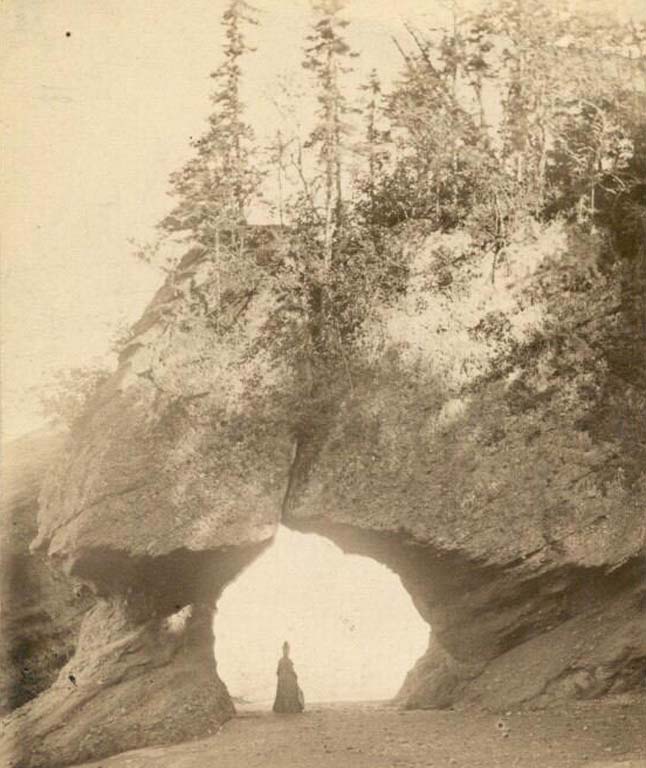 Black and white photograph of arched rock formation with woman in long dress standing in open arch on beach