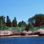 Colour photograph of water, reddish rock shoreline and trees