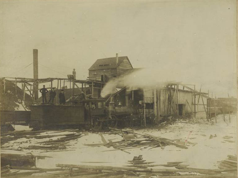 Black and white photograph of wood building with three men and a trail locomotive