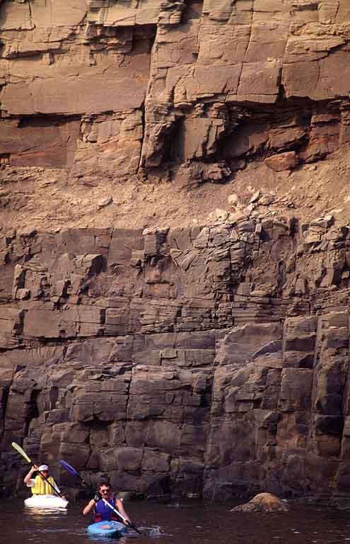 Colour photograph of a brown rock cliff on the ocean with two people in kayaks